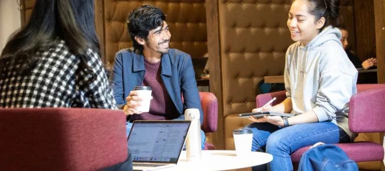 three students sat at a table talking with laptops and notepads