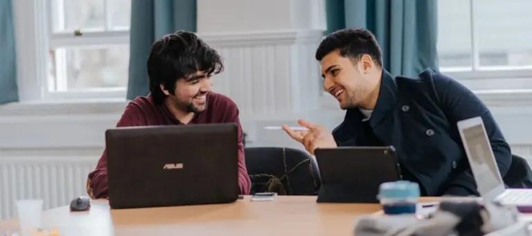 two students talking at a table with a laptop